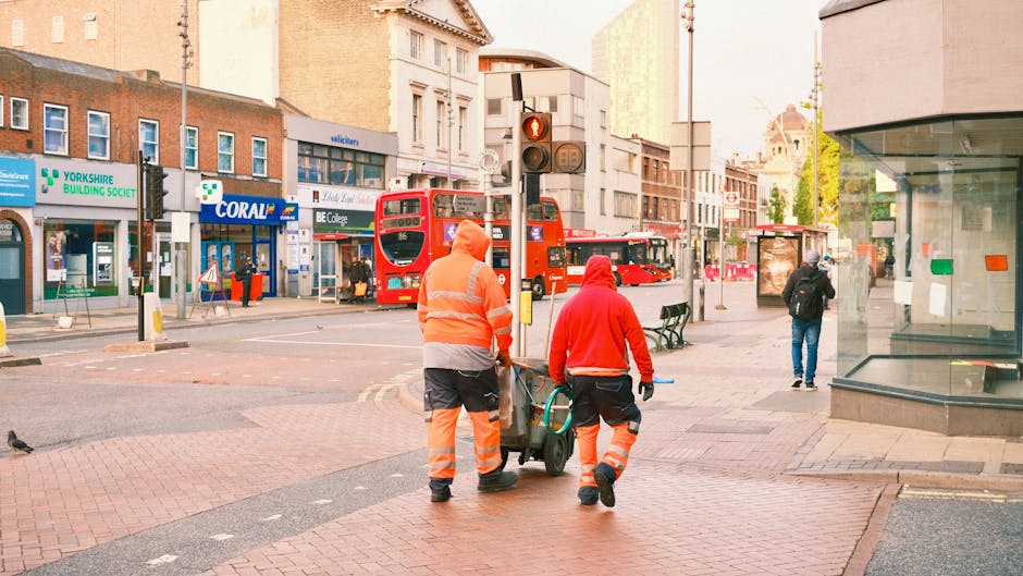 Two street cleaners in orange high-visibility uniforms and jackets are walking along a busy urban street near Colliers Wood Station, with one pushing a cart. The scene includes a red double-decker bus, pedestrians, and various storefronts with signage for local businesses like Yorkshire Building Society, Coral, and B.E. College. The area features brick and glass buildings, a traffic signal, benches, and a bus stop. The street appears clean with well-maintained pavements and minimal litter, indicating effective maintenance and cleanliness. The photo is taken during daylight with natural lighting, capturing a typical city street environment. Cleaner Colliers Wood specializes in surface cleaning, deep cleaning, and sanitisation services for residential and commercial spaces in the Colliers Wood area.