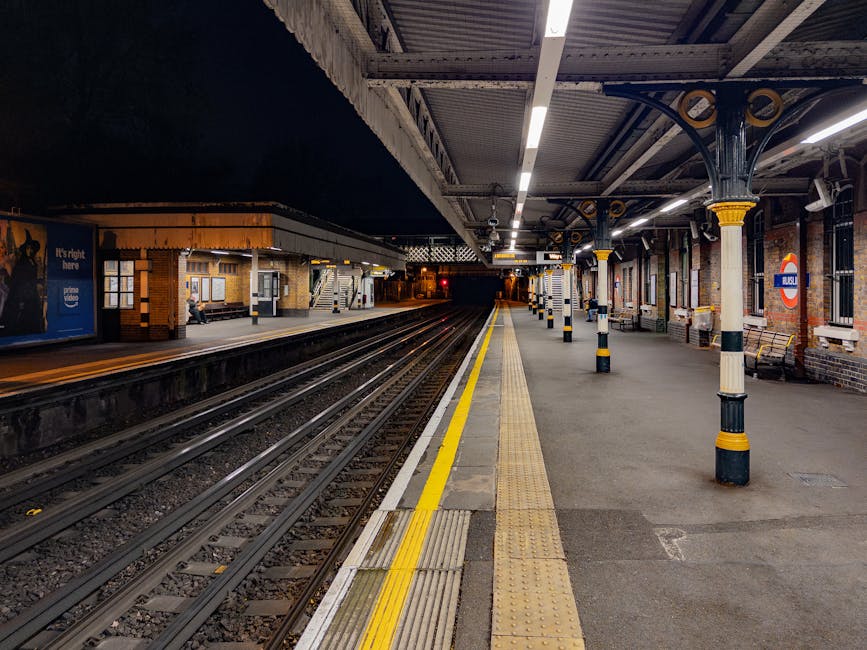 A clean and well-lit underground railway platform at night, with polished stone flooring adjacent to railway tracks. The platform features concrete and brick walls with window openings, metallic support pillars with black and yellow safety markings, and overhead lighting illuminating the area. There are benches, signage, and a station sign with the London Underground logo visible on one pillar. The surface area appears tidy, free of litter, with reflective yellow safety lines along the platform edge. This setting showcases the importance of routine cleaning and maintenance in railway stations, as provided by Cleaner Colliers Wood's deep cleaning services near Colliers Wood Station, ensuring hygiene and safety for passengers.