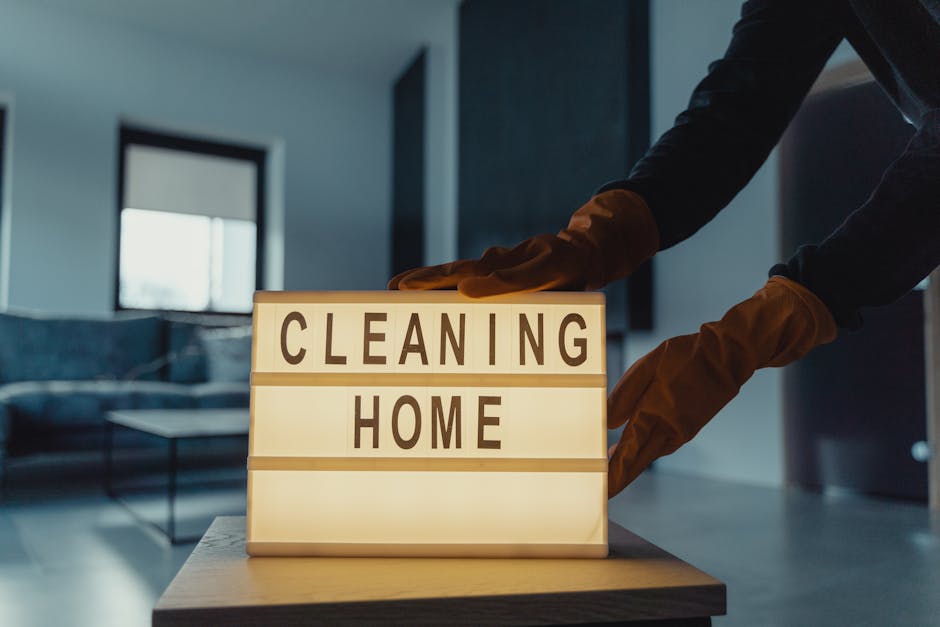 A person wearing brown cleaning gloves is positioning a small illuminated sign that reads 'CLEANING HOME' on a wooden table in a modern living room. The room features a glass coffee table with a metal